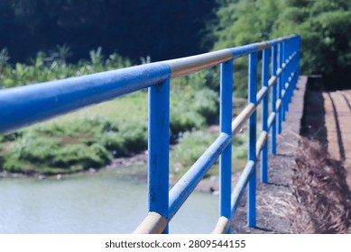 Blue railings line a rustic path beside a stream. Tall trees and greenery surround the walkway. Sunlight casts long shadows across the dirt trail
