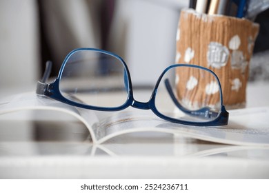 Blue reading glasses placed on an open book in a study setting with a pencil holder. Ideal for themes of education, reading, and focus. - Powered by Shutterstock