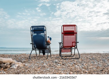 Blue and red chairs on pebbles beach at seaside in southern Italy, one man from behind sitting and relaxing with a smartphone and enjoyngthe sea at fall