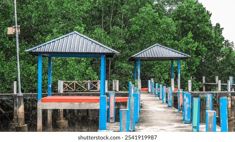 A blue and red pavilion sits on a pier next to a dock. The pavilion is open and has a bench underneath it. The dock is surrounded by trees and the water - Powered by Shutterstock