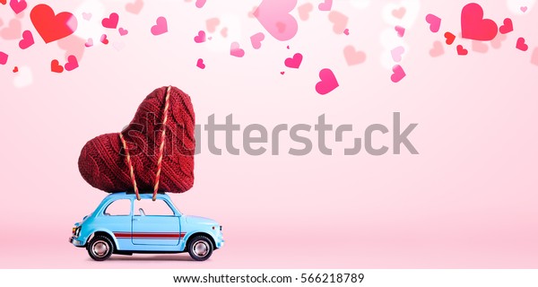 Blue retro toy car delivering\
craft heart for Valentine\'s day on pink background with\
confetti