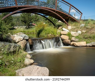 The Blue River Flowing Through Steamboat Springs.  Green Bushes And Trees Along With Blue Water. Bridgework Over A Waterfall.