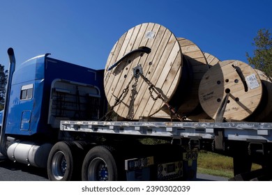 a blue semi truck carries large wooden spools secured by metal chains on its open back trailer bed on a bright sunny day on the highway