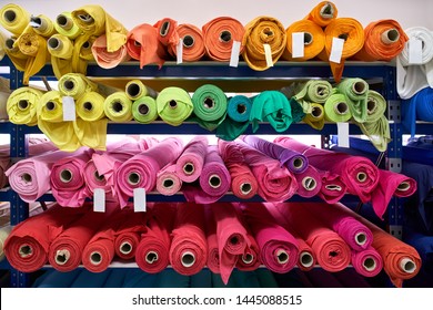 Blue Shelves With Bright Multicolored Fabric Rolls On The Light Wall Background Inside A Warehouse. Closeup Horizontal Photo.