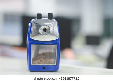 A blue and silver pencil sharpener sits on a white surface. The sharpener has a metal blade frame on top with two black buttons. - Powered by Shutterstock