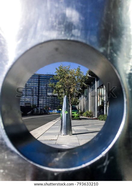Blue\
skies and green trees reflected in a metal post and further views\
down a deserted street visible through an\
aperture