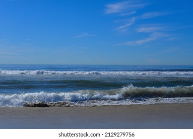Blue skies with some light clouds above crashing waves along the Cardiff California beach. The horizon is centered allowing copy space in the sky. Contrasting beach crashing waves in the foreground.