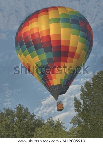 Blue sky and multicolored hot air balloon with wicker basket and passengers rising above treetops shortly after liftoff. Flames visible from the burner unit.