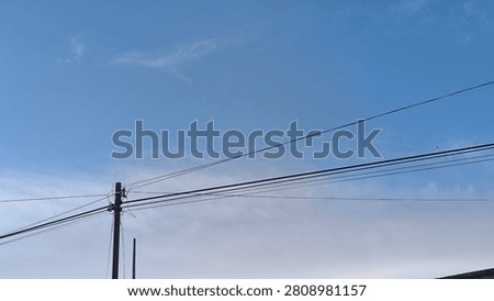 Blue sky with soft clouds above power lines. Wooden pole supports multiple cables stretching across frame. Daylight scene shows urban infrastructure and open atmosphere. Background provides clear space.