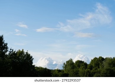 Blue sky with thin white clouds above a green forest canopy. Natural landscape with open sky, treetops, and summer weather conditions.