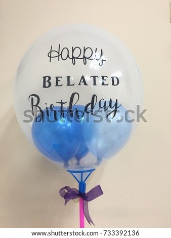 Blue small balloons inside transparent ballon with “Happy Belated Birthday” on it stick on pink stick and purple ribbon