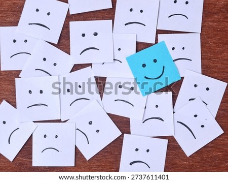 Blue smiling face adhesive note among many white sad faces hand drawn on wooden background. Positivity Concept for optimism, mental health awareness and staying positive