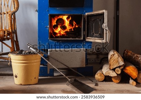 A blue solid fuel boiler with an open door reveals burning logs inside, emitting a warm glow. Stack of chopped wood is arranged on the floor. There are pieces of coal in a yellow metal bucket.