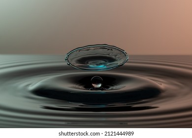 Blue Splash Of Water Hovering Above A Single Water Drop Which Is Resting On Dark Water