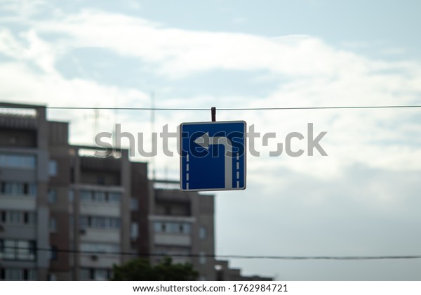 A blue\
square road sign hanging on a cable with a white arrow pointing to\
the left. With blurry\
background.