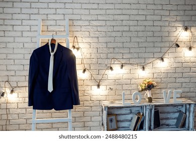 A blue suit and white tie hang on a white ladder against a brick wall decorated with string lights. A bouquet of flowers sits on a shelf with the word LOVE spelled out. - Powered by Shutterstock