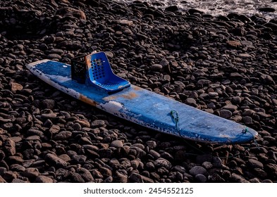 A blue surfboard with a chair on it is sitting on a rocky beach. The chair is missing and the surfboard is old and rusted. Scene is somewhat sad and lonely - Powered by Shutterstock