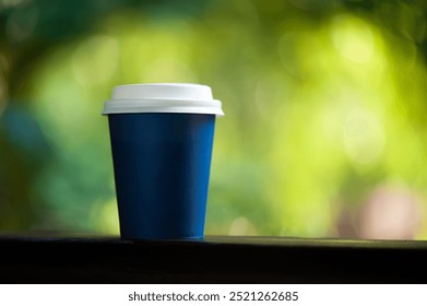 A blue takeaway coffee cup with a white lid placed outdoors, with a blurred green nature background - Powered by Shutterstock