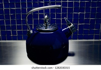 A Blue Tea Kettle Sitting On A Stainless Steel Counter With A Unique Blue Glass Tile Background