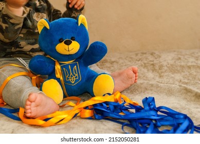 Blue teddy bear with trident on the abdomen in the hands of child. Baby with blue teddy bear in his arms sits next to blue and yellow satin ribbons. Ukrainian child with his toy. Conflict in Ukraine