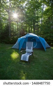 Blue Tent On A Camping Site With An Empty Relaxing Chair In Front Of It With A Sun Flare Through The Forest