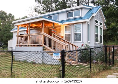 Blue tiny house with wooden porch surrounded by a black chain link fence. Warm glowing porch lights with a pumpkin welcome sign hanging from porch.
