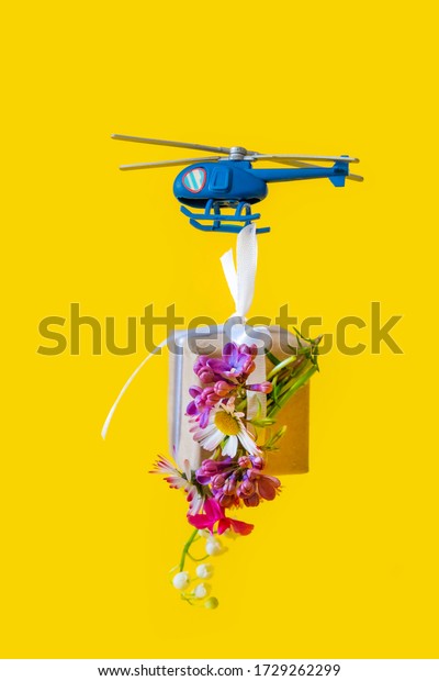 Blue Toy Helicopter Delivers Gift Paper box with\
flowers on the Yellow Background. Mother\'s day, Valentine\'s day,\
Father\'s day, Birthday\
concept.