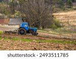 A blue tractor plows a field in a rural landscape. The tractor is moving to the right, leaving behind a freshly plowed furrow. The field is surrounded by trees and hills.