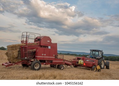 A blue tractor pulling a red hay bailer is parked for the night in a mowed hay field. beautiful sly dominates the scene with puffy white clouds, A mountian range is in the background.