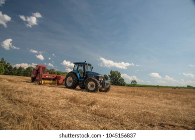 A blue tractor pulling a red hay bailer is parked in a mowed hay field awaiting the next day of work. The sky is a dramitic blue with small puffy white clouds.
