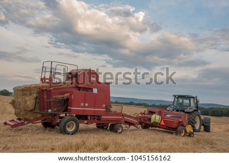 A blue tractor pulling a red hay bailer is parked for the night in a mowed hay field. beautiful sly dominates the scene with puffy white clouds, A mountian range is in the background.