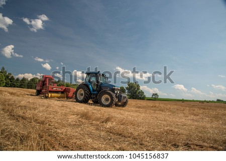 A blue tractor pulling a red hay bailer is parked in a mowed hay field awaiting the next day of work. The sky is a dramitic blue with small puffy white clouds.