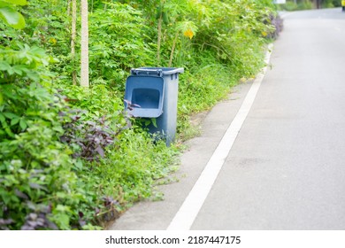 The Blue Trash Bin Beside The Asphalt Road Has A Lot Of Trees.