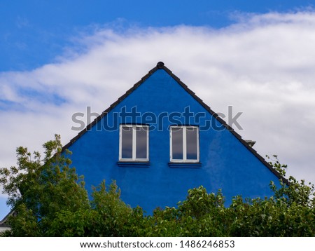 Blue triangle bulding with two windows and trees on cloudy sky background