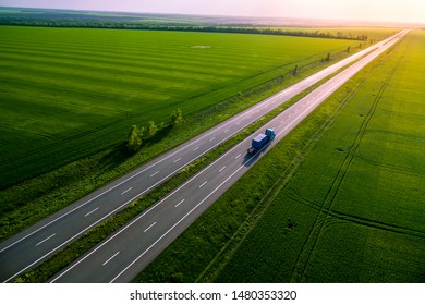 Blue Truck On The Higthway Sunset. Cargo Delivery Driving On Asphalt Road Along The Green Fields. Seen From The Air. Aerial View Landscape. Drone 