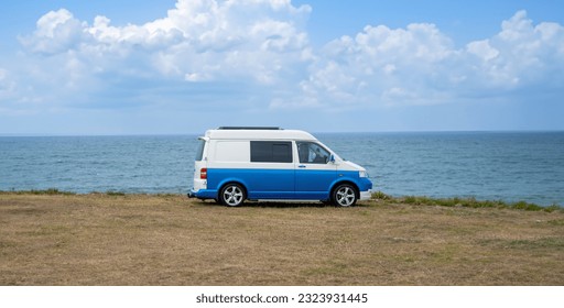 Blue vintage camper van parked on a grassy plain with the Atlantic Ocean in the background under a semi-cloudy sky lit by the early morning sun.