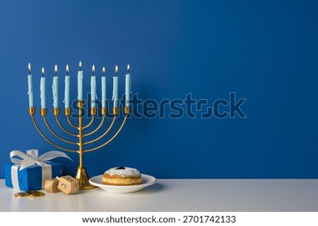 Blue wall backdrop frames a golden menorah with lit candles, a wrapped gift, dreidel, coins, and a donut on a plate, capturing warm festive light and Hanukkah spirit