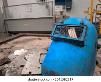 A blue welding helmet placed on a dusty, metal workbench in a workshop, showing marks of use and protective wear - Powered by Shutterstock