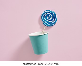 Blue and white colored swirl round candy lollipop in a paper cup on pink background.