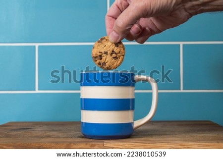 Blue and white cup of tea with a hand dunking a chocolate chip biscuit cookie. Blue and white cup isolated against a blue tiled background. Mug of hot drink with a sweet treat. 