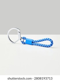 A blue and white patterned rope keychain with a silver metal ring rests on a white surface against a neutral light grey studio background, studio.