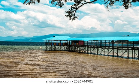 A blue and white pier is on the water. The pier is surrounded by a sandy beach. The sky is cloudy and the water is calm - Powered by Shutterstock