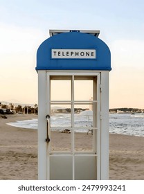 A blue and white telephone booth on a beach, with the sea and buildings in the background.