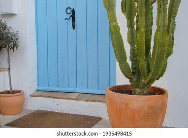 A Blue Wooden Door With Black Old Handle. A Door Mat Below A Tiled Step In This Welcoming, Homely, Rustic Entrance. A Cactus In A Pot On The Right And Olive On The Left For European Beach Holiday Feel