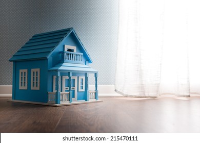 Blue wooden model house next to a window with curtain on wooden floor.