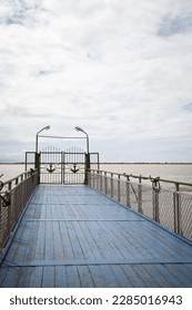 Blue wooden pier with railing. River dock ending with gates decorated with anchors. Horizon and cloudy sky on the background. Quiet, peaceful beautiful view