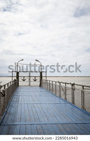 Blue wooden pier with railing. River dock ending with gates decorated with anchors. Horizon and cloudy sky on the background. Quiet, peaceful beautiful view