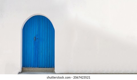 Blue wooden vintage closed door with arch on an empty whitewashed wall background. Cyclades Greek island architecture, blank copy space card template advertise.