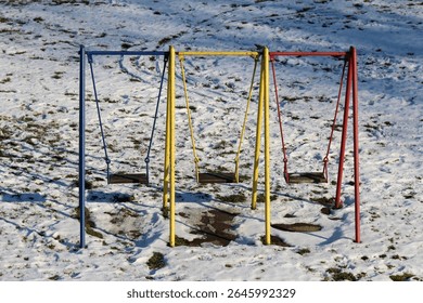 Blue, yellow, and red metal swingset with three empty swings stands on a playground covered in patchy snow, with visible footprints and grass beneath the thawing surface