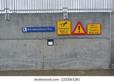 Blue, yellow, and red warning signs on a grey concrete wall with steel railing in Iceland